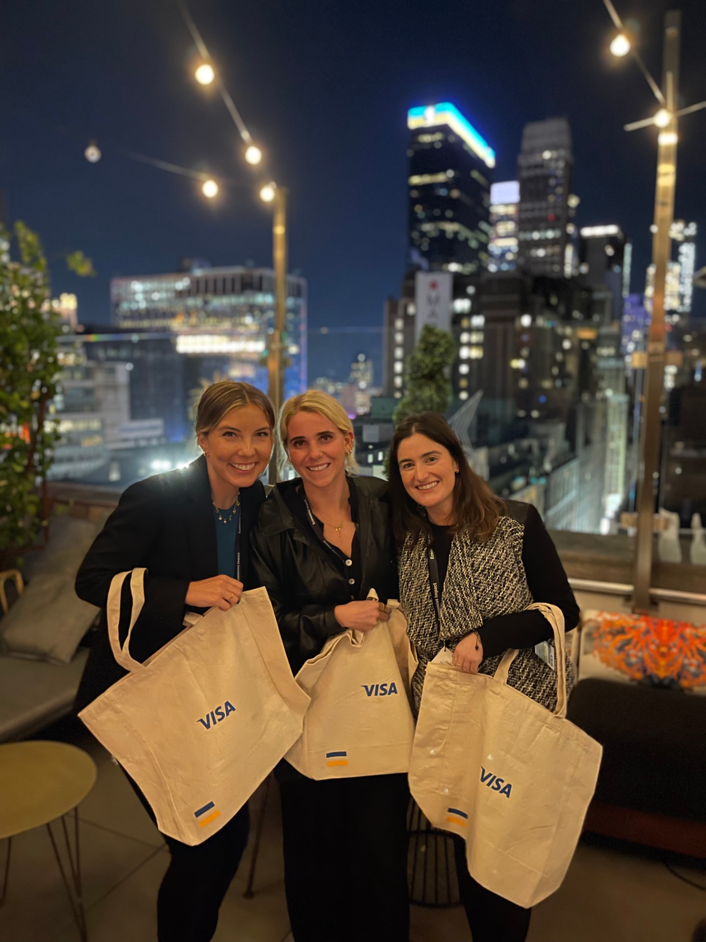 Three guests holding Visa canvas tote bags on the rooftop with the city skyline behind them