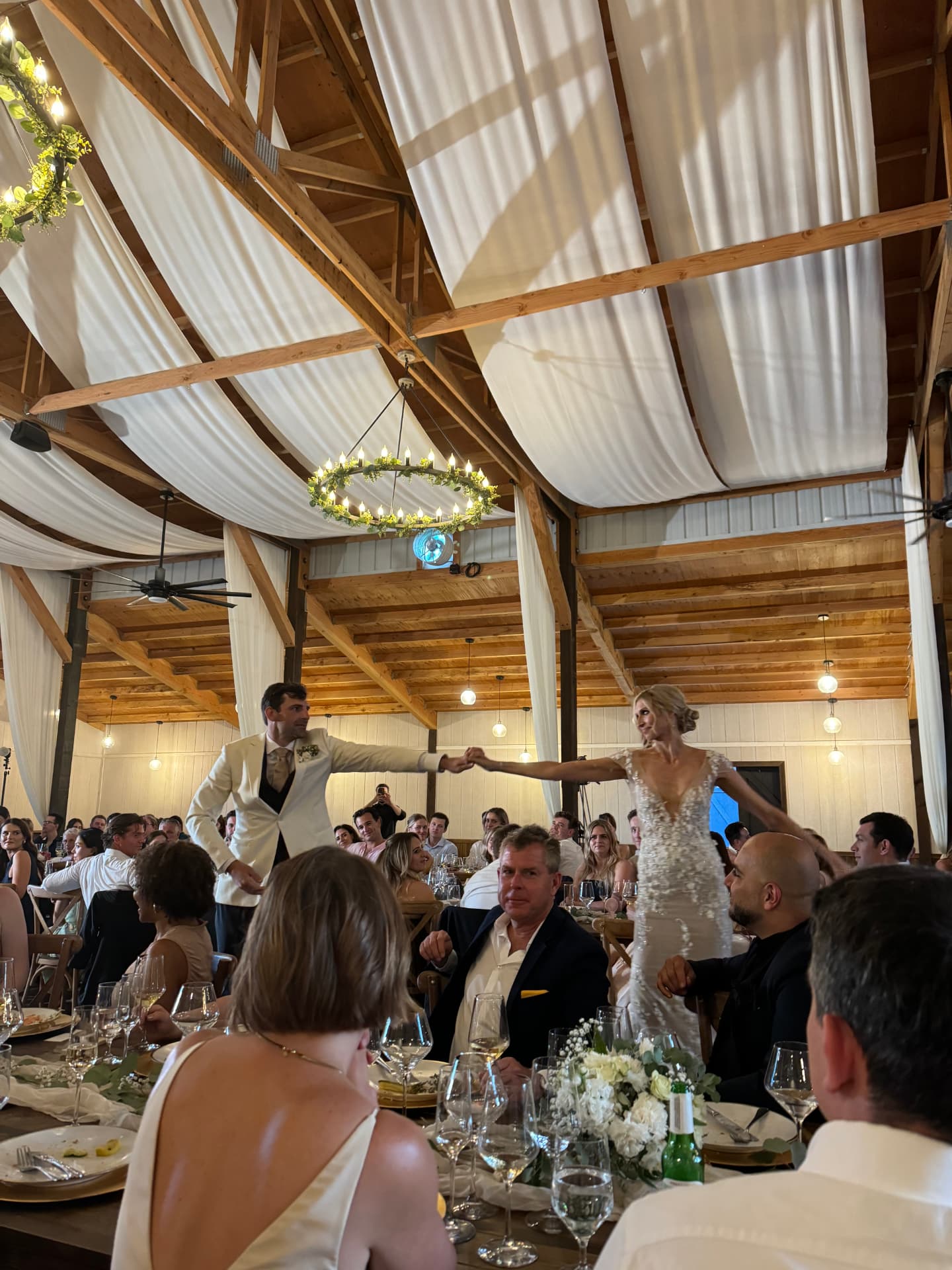 Bride and groom hand in hand during their first dance under a floral chandelier in the barn