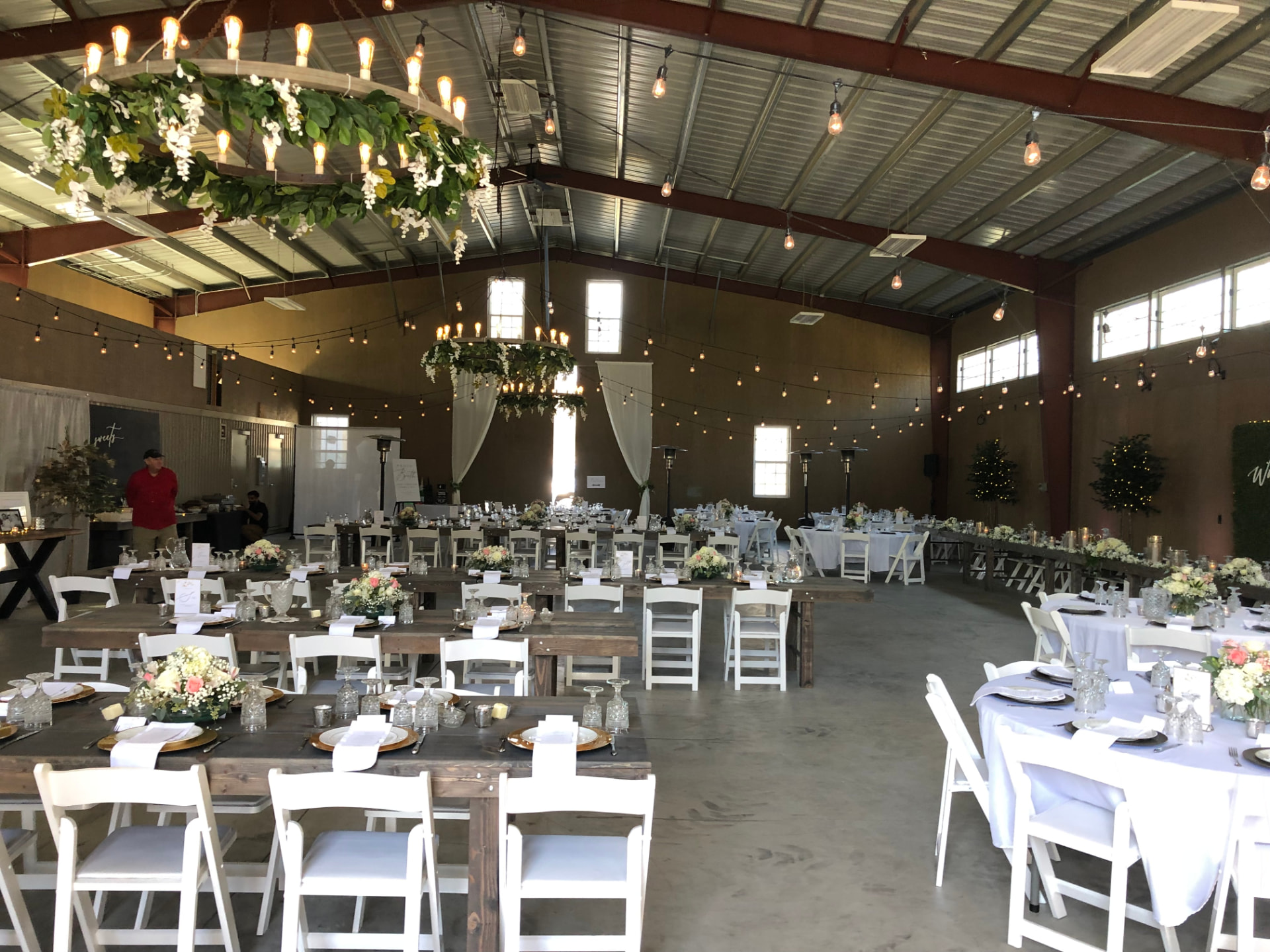 Barn reception set with long farm tables, greenery chandeliers and string lights before guests arrive