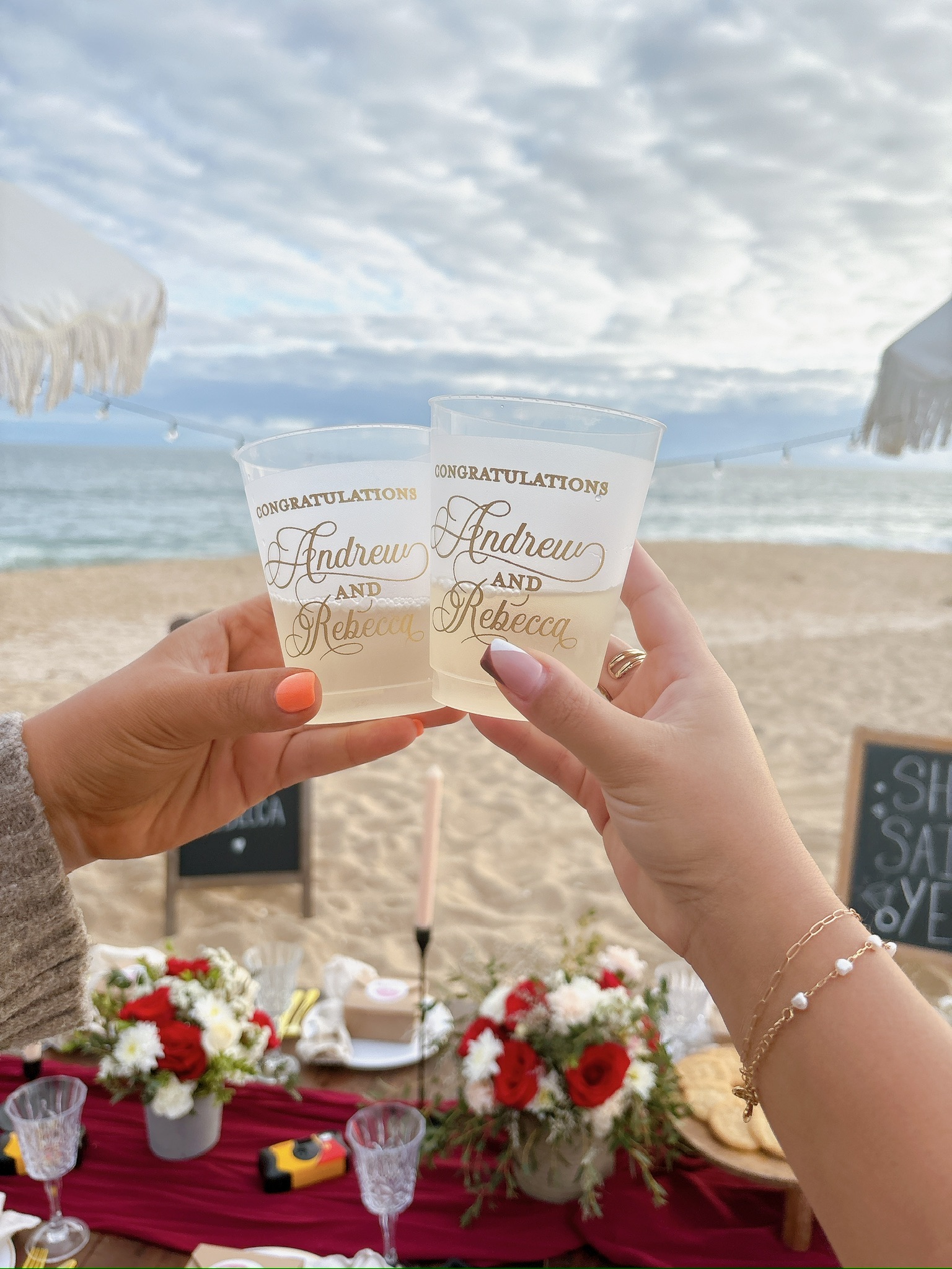 Beach engagement party - two hands toasting with custom congratulations cups at beachside celebration