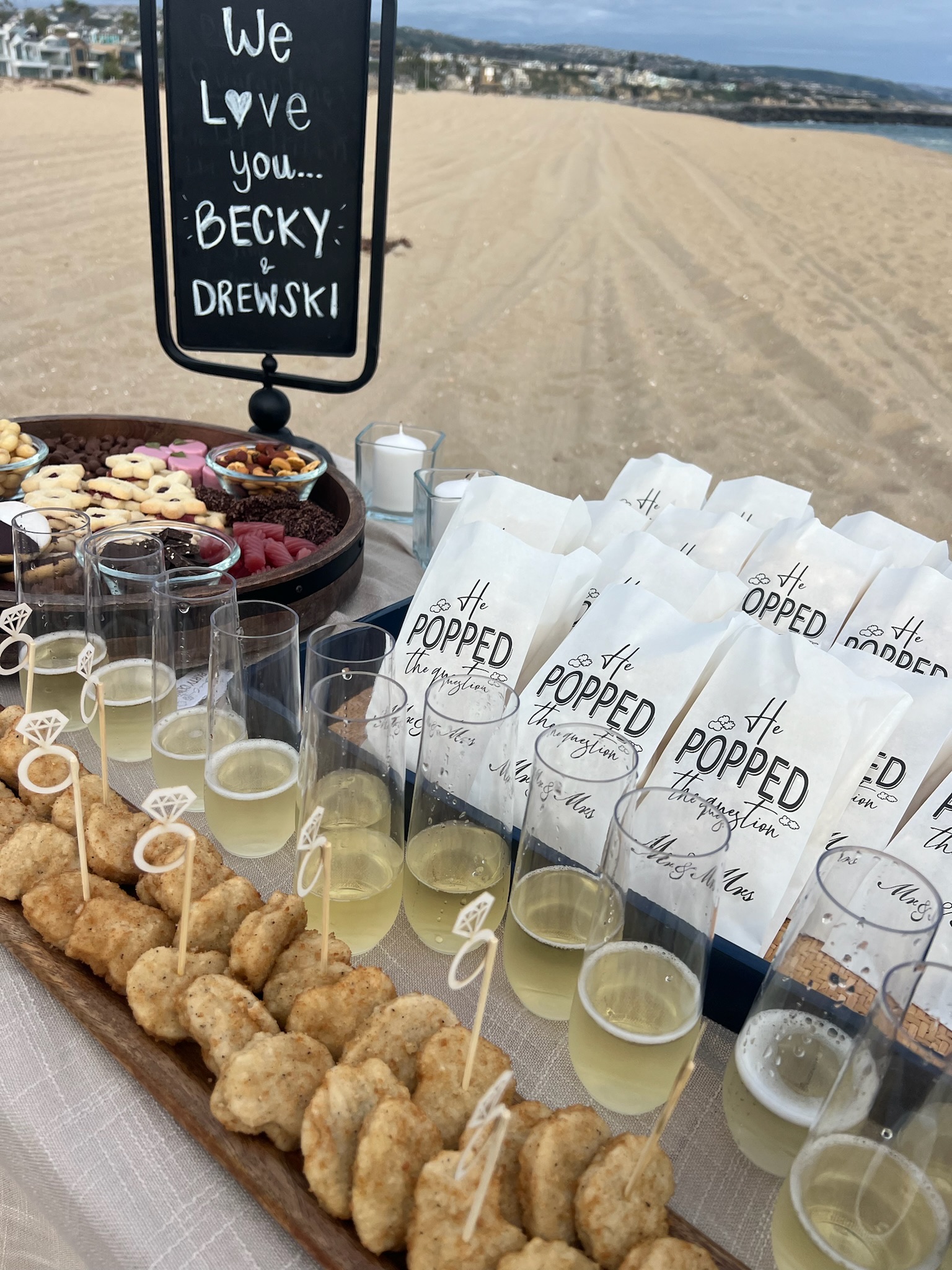 Beach engagement party - setup with chalkboard sign, champagne glasses, and 'He POPPED the question' favor bags