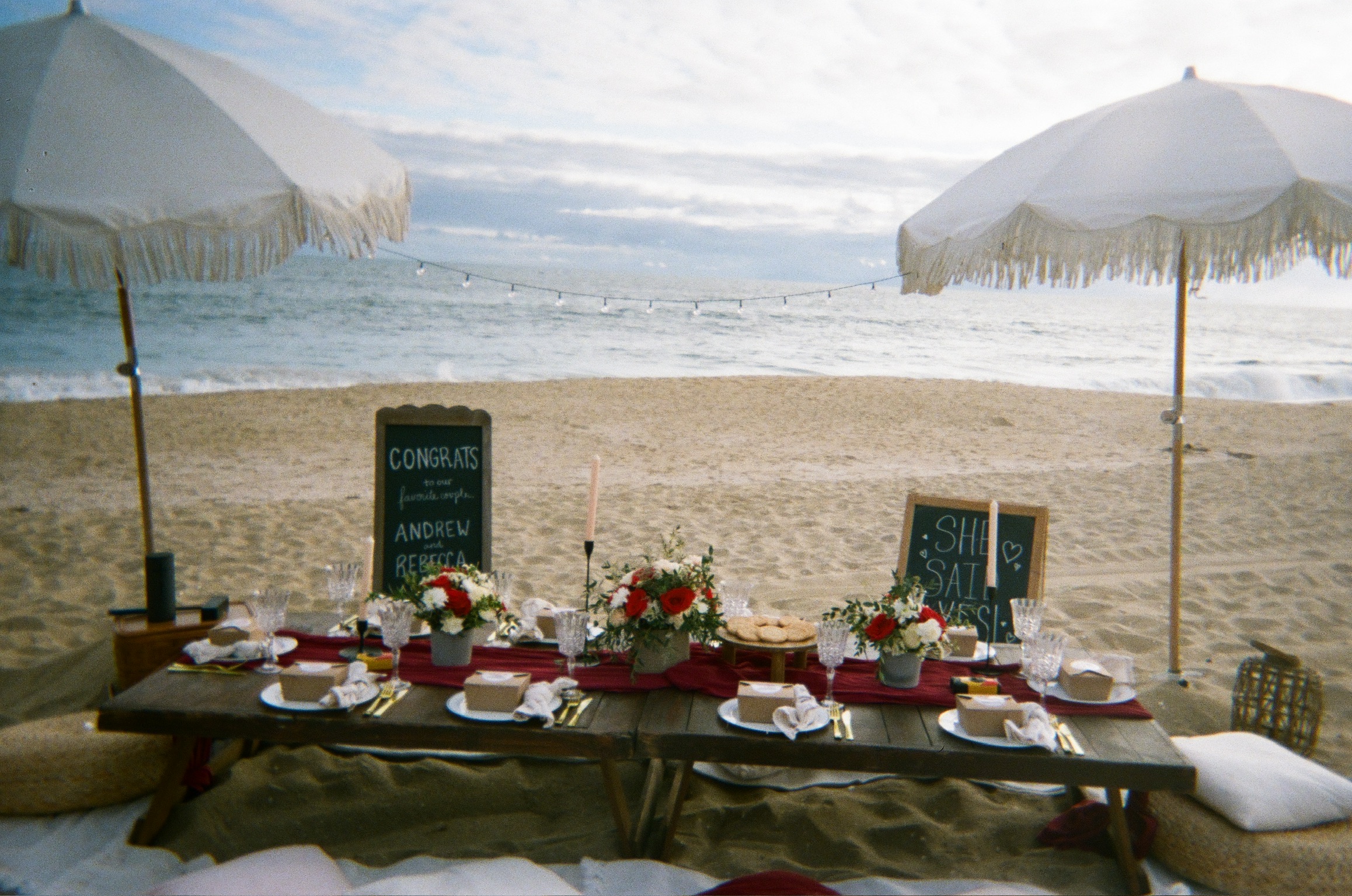 Beach engagement party - wide shot of beachside celebration with white umbrellas and 'SHE SAID YES!' sign