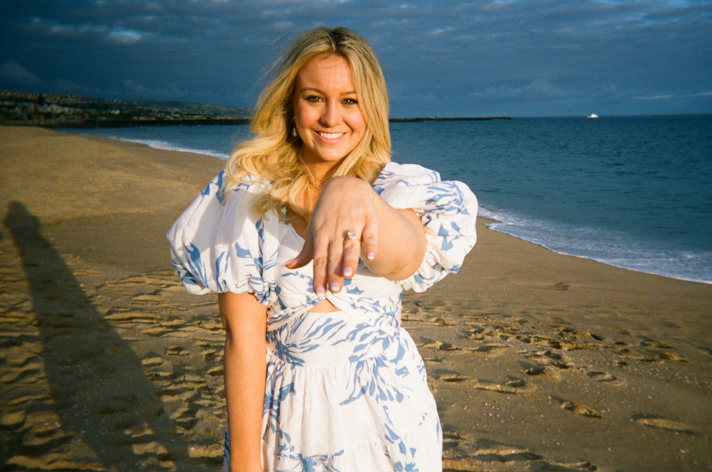 Beach engagement party - portrait of bride-to-be in floral dress showing engagement ring with ocean backdrop